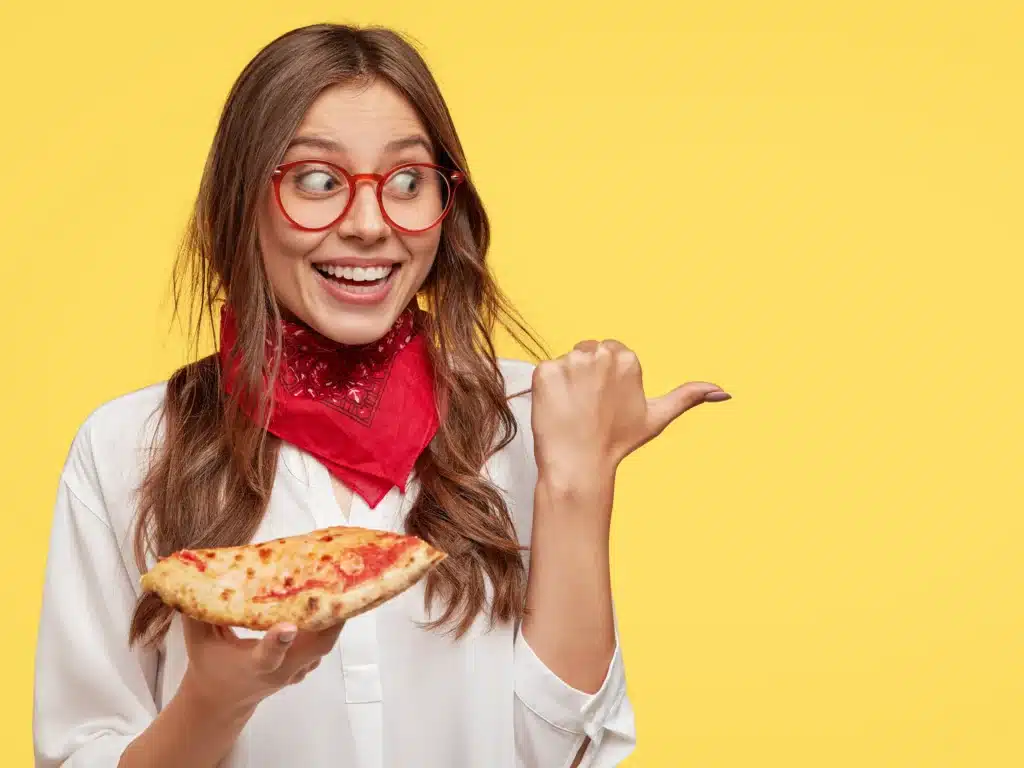 Iloinen nainen pizza kädessä soittaa sormella ja katsoo sivuun.